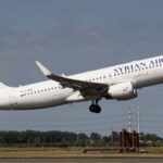 Syrian Air Airbus A320 climbs after take-off with landing gear extended, flying above a runway under a clear blue-grey sky.