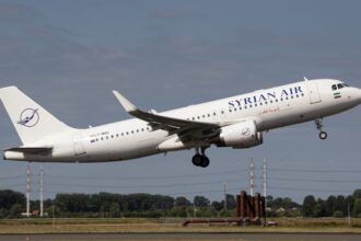 Syrian Air Airbus A320 climbs after take-off with landing gear extended, flying above a runway under a clear blue-grey sky.