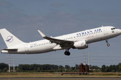 Syrian Air Airbus A320 climbs after take-off with landing gear extended, flying above a runway under a clear blue-grey sky.