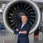 A photo of Michalis Kouveliotis in a blue suit, who is stood in front of a big jet engine with his arms crossed, smiling. The building surrounding him is white with a very shiny and reflective floor.