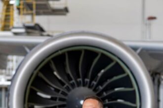 A photo of Michalis Kouveliotis in a blue suit, who is stood in front of a big jet engine with his arms crossed, smiling. The building surrounding him is white with a very shiny and reflective floor.