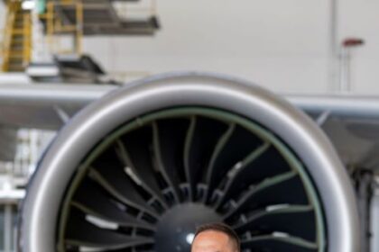 A photo of Michalis Kouveliotis in a blue suit, who is stood in front of a big jet engine with his arms crossed, smiling. The building surrounding him is white with a very shiny and reflective floor.