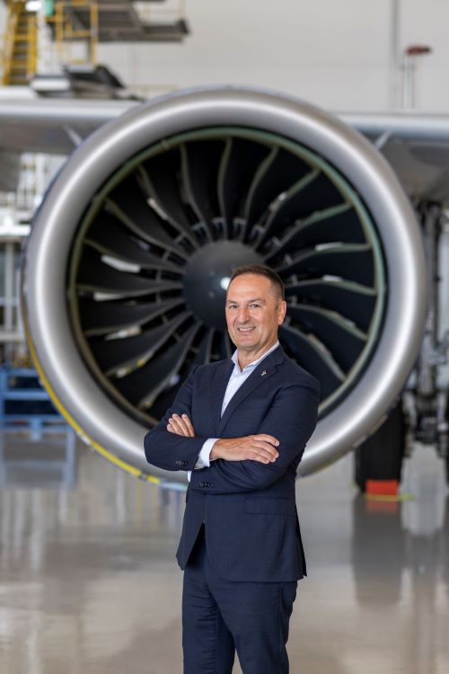 A photo of Michalis Kouveliotis in a blue suit, who is stood in front of a big jet engine with his arms crossed, smiling. The building surrounding him is white with a very shiny and reflective floor.