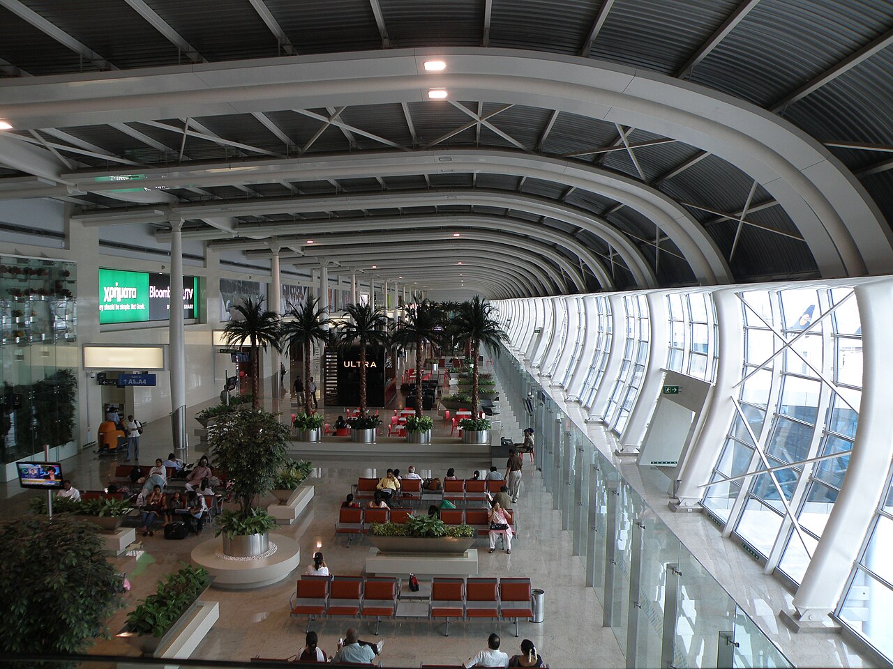An image of the interior of Mumbai International Airport Terminal 1, where construction is set to take place in January 2027. The structure of the building is white and shaped geometrically, with many windows lined up along the airport's tarmac. Many plants and greenery can be seen inside the terminal, and the building has a general cleanliness about it.