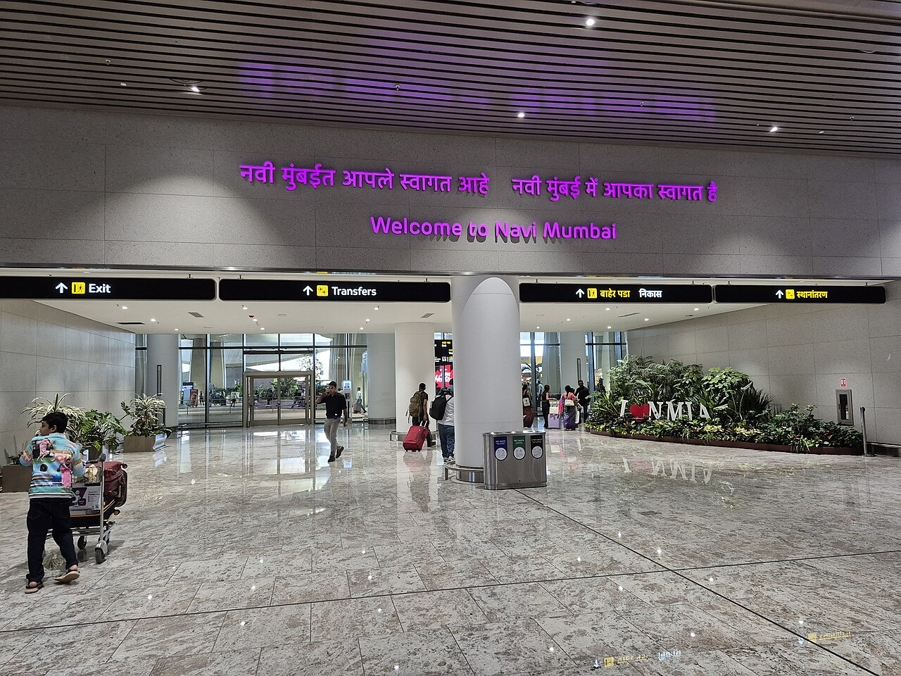The inside entrance of Navi Mumbai International Airport, in which the interior is designed with light sandy colours and some woody accents. The lighting is bright and cold, with the entrance featuring the sign 'Welcome to Navi Mumbai' in bright purple.