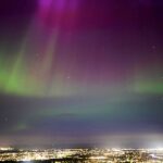 Image shows the Northern Lights above the city of Edinburgh viewed from Holyrood park. City lights can be seen in the foreground with the night sky a range of rainbow colours