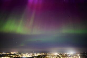 Image shows the Northern Lights above the city of Edinburgh viewed from Holyrood park. City lights can be seen in the foreground with the night sky a range of rainbow colours