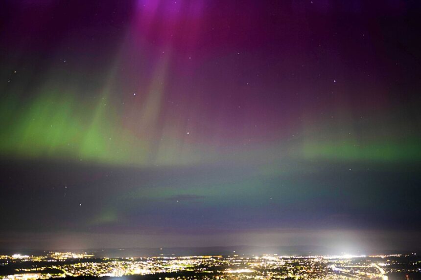 Image shows the Northern Lights above the city of Edinburgh viewed from Holyrood park. City lights can be seen in the foreground with the night sky a range of rainbow colours