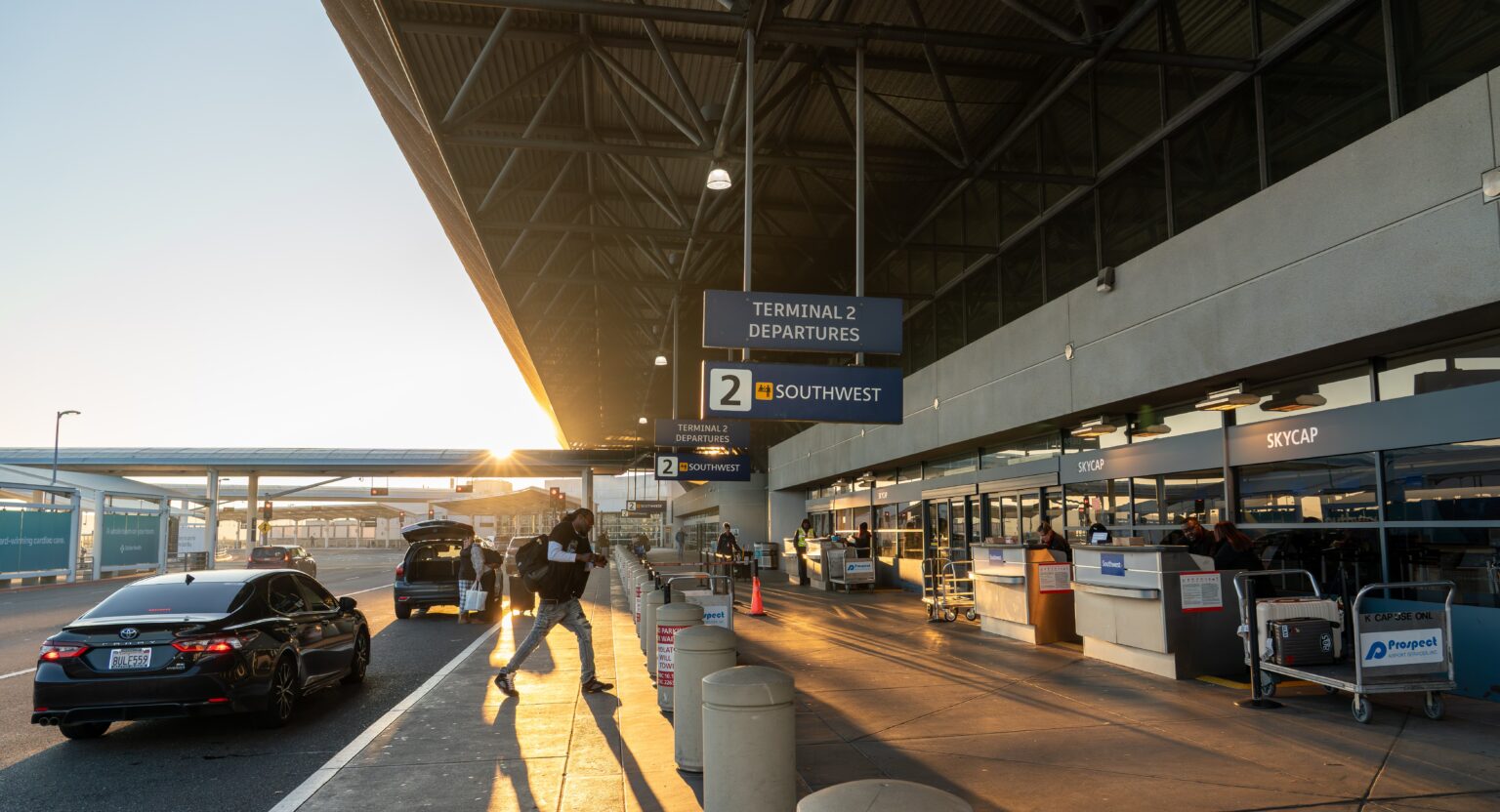 Photograph of an airport Terminal 2 departures curb at sunrise, showing passenger drop-off activity and travelers entering terminal.
