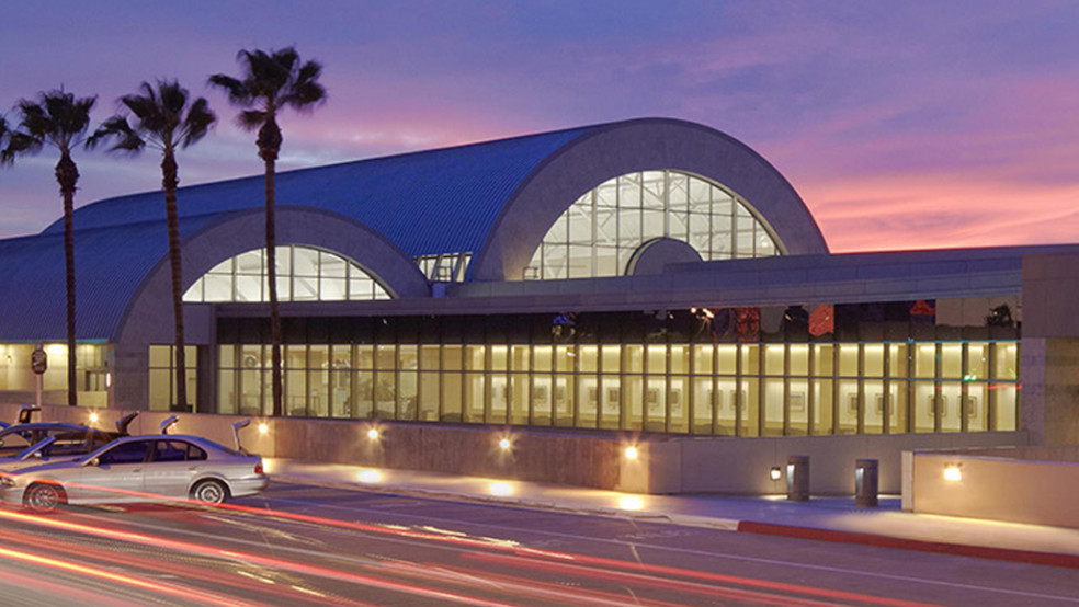 John Wayne Airport terminal exterior at dusk, featuring blue curved roofs, large arched glass windows, palm trees, and illuminated entrance. Long-exposure traffic light trails and parked cars line roadway, highlighting busy transportation setting and dramatic purple-pink sunset.