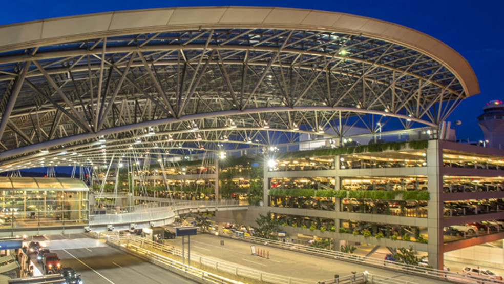 Photograph shows a modern airport terminal at dusk, featuring a sweeping steel-and-glass canopy and brightly lit interior. Elevated roadways, vehicle lanes, landscaped terraces, multilevel parking, and an illuminated control tower highlight airport access and infrastructure.