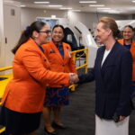 Crew pictured smiling in bright orange blazers and shaking hands with Qantas facilities at the back.
