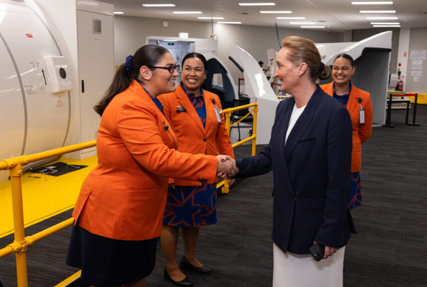 Crew pictured smiling in bright orange blazers and shaking hands with Qantas facilities at the back.