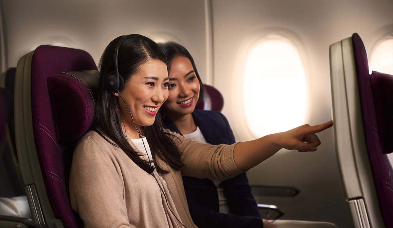 Photograph depicting Qatar Airways Economy Class cabin with in-flight entertainment, depicting two women seated together with one of them gesturing towards the in-flight entertainment screen.