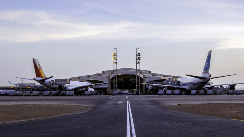 Commercial airplanes parked at airport gates in front of a terminal building, viewed from the airport taxiway at dusk.