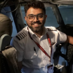 Image shows Smit Machcchar in the cockpit of a plane, smiling at the camera. He has dark hair and a beard, wearing a pilot's uniform and glasses.