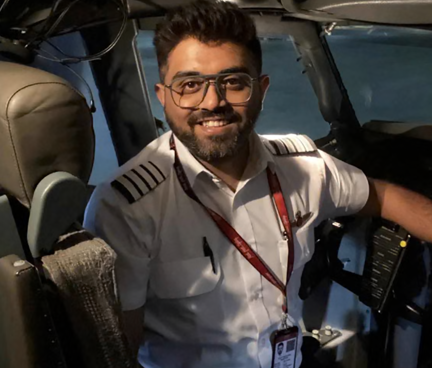 Image shows Smit Machcchar in the cockpit of a plane, smiling at the camera. He has dark hair and a beard, wearing a pilot's uniform and glasses.