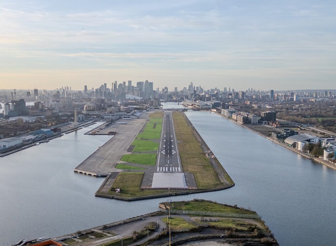 Image shows London City Airport from an aerial view, looking down the runway. The London skyline is visible on the horizon.