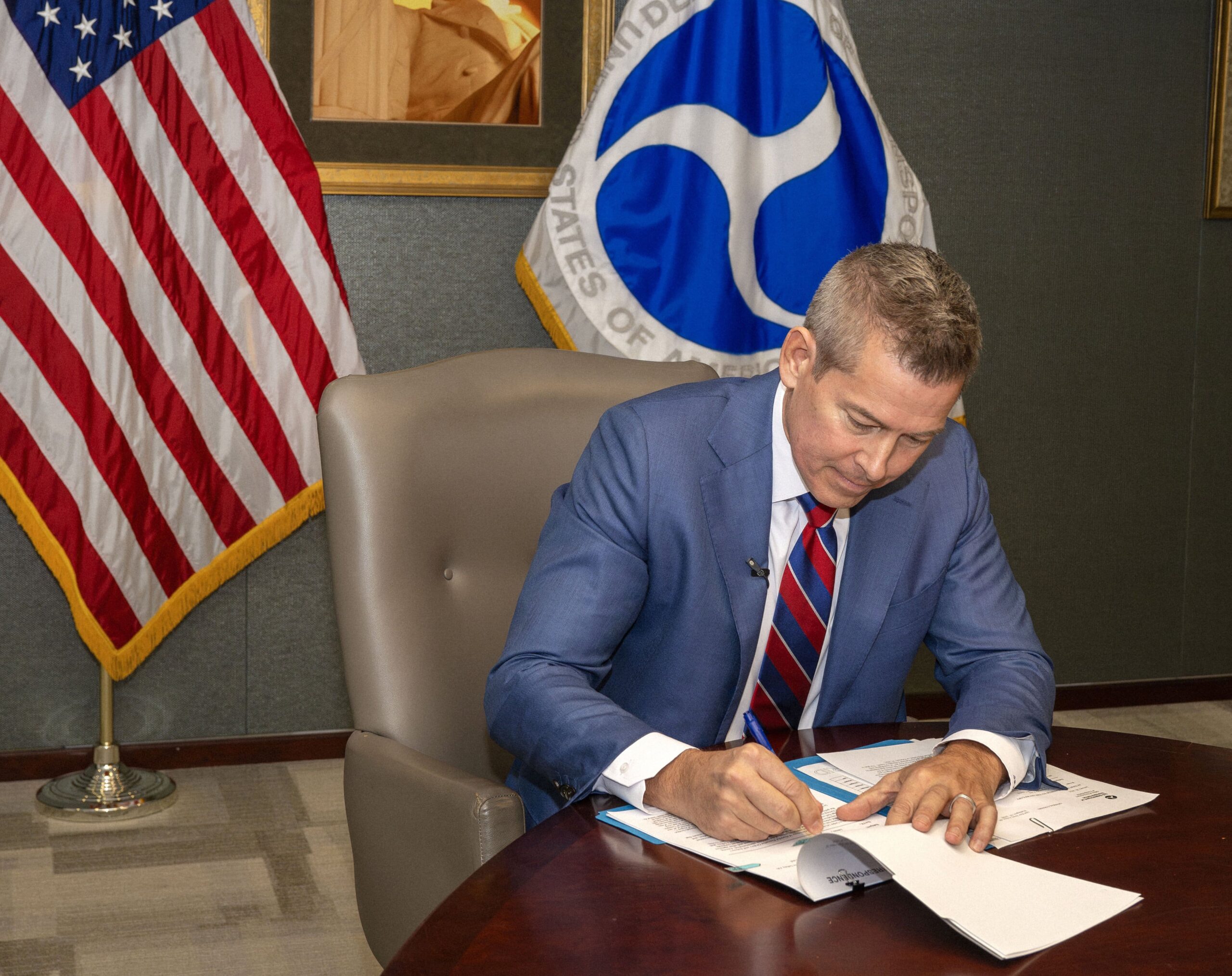 Man in a blue suit and striped tie signs a document at a round table, with a US flag and a Department of Transportation flag behind him.