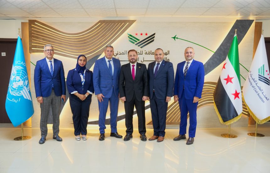 Six officials pose between ICAO and Syrian civil aviation flags in a government office, standing before a wall display with aviation-themed graphics.