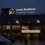 Leeds Bradford Airport at night in the middle of a light snowstorm. The sign shines brightly announcing it's LBA. The sky is nearing pitch black and warm lights glow from within the airport.