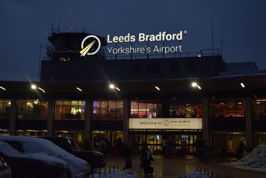 Leeds Bradford Airport at night in the middle of a light snowstorm. The sign shines brightly announcing it's LBA. The sky is nearing pitch black and warm lights glow from within the airport.