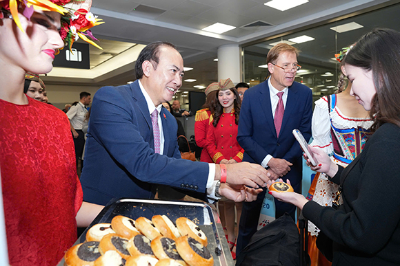 Two men in suits take pastries from a tray in Prague airport terminal while women in traditional dress stand nearby.