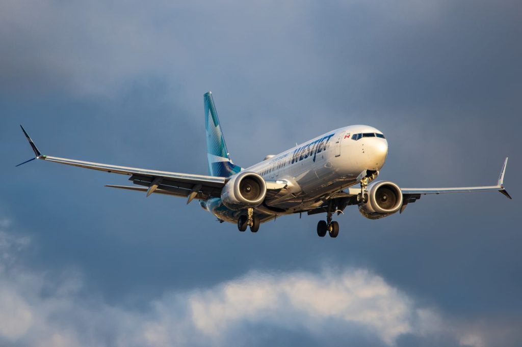 A WestJet passenger jet flying high above, with a bright sky and scattered clouds surrounding it.