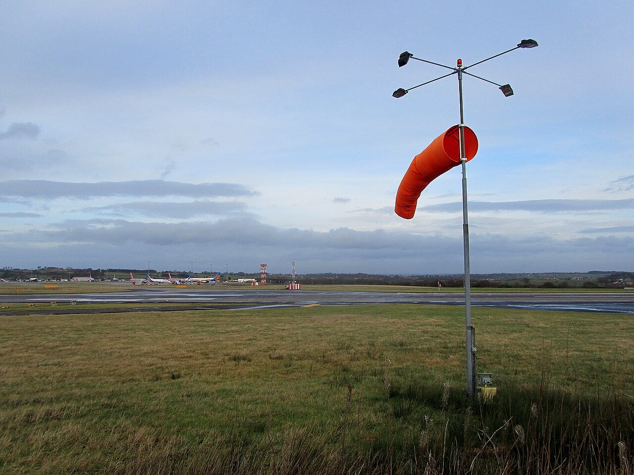A red windsock by Leeds Bradford Airport on a cloudy, stormy day is being blown about. The sky is a grey-blue with clouds slowly rolling in from the distance. In the foreground, the grass is a muddy green colour and behind it is the runway containing stationary aircraft.
