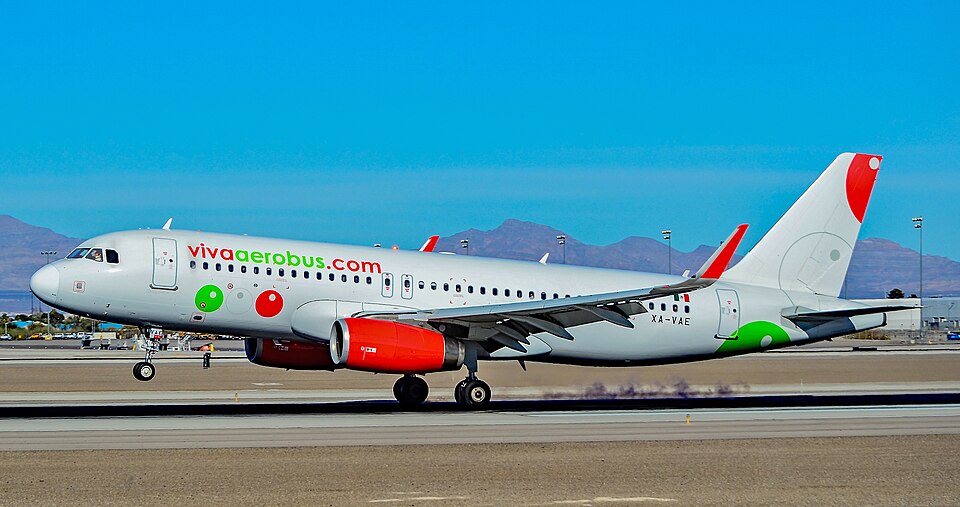Grey jet with red engines and green and red dots on its fuselage, landing gear touching the runway, with desert mountains behind under a clear blue sky.
