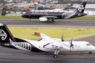 Two Air New Zealand aircraft on airport taxiways, including a De Havilland Q300 turboprop in the foreground and an Airbus narrowbody aircraft in the background, with airport buildings and residential areas visible beyond the airfield.