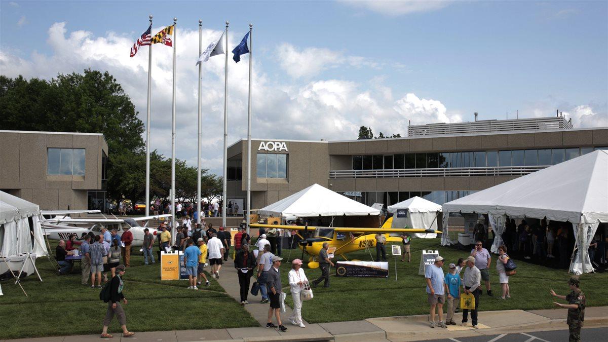 Image depicting the AOPA headquarters. People are seen walking between gazebos under four flags flying. A yellow aircraft stands between them.