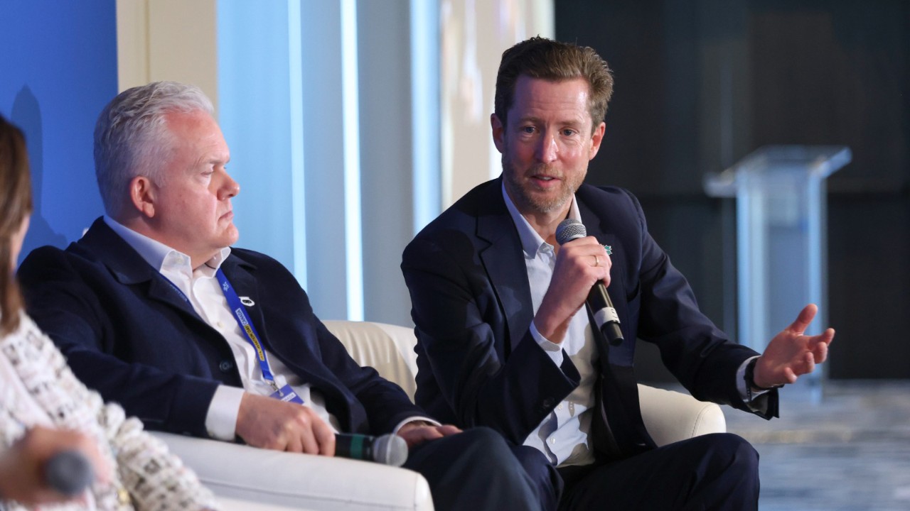 Alexis von Hoensbroech, WestJet CEO, is sat down holding a microphone, mid speech. He is sat next to two other people, both looking at him.