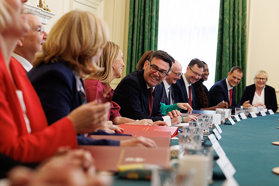 Several people are sat along a long table, all facing away from the camera, with Prime Minister Andy Burnham sat in the middle of the group turned to face them, smiling.