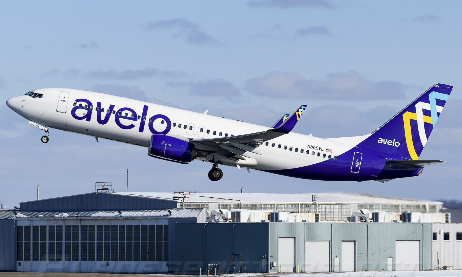 An Avelo Airlines B737 having just taken off, ascending into the air. The top of a building can be seen in the background along with a blue sky.