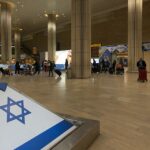 The inside of Ben Gurion International Airport (TLV) in Israel. The Israeli flag is at the forefront of the image. In the background, there are groups of people with luggage walking through the terminal.