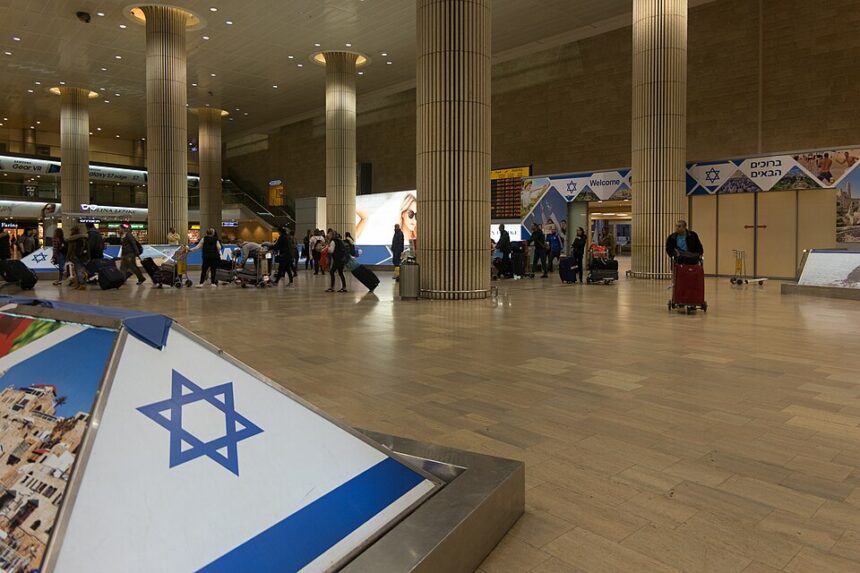 The inside of Ben Gurion International Airport (TLV) in Israel. The Israeli flag is at the forefront of the image. In the background, there are groups of people with luggage walking through the terminal.