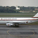 A Biman Bangladesh Airlines Airbus plane that has come to a a stop on a concrete runway. There is green grass and foliage in the background of the photo, alongside some buildings. The sky is grey and cloudy.