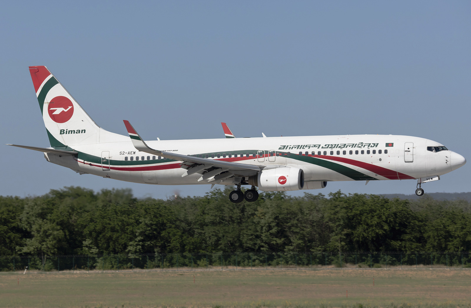A Biman Bangladesh Airlines B737 approaching landing. The ground and trees can be seen in the background, as well as the blue sky.