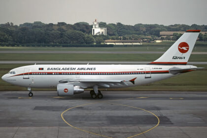 A Biman Bangladesh Airlines Airbus plane that has come to a a stop on a concrete runway. There is green grass and foliage in the background of the photo, alongside some buildings. The sky is grey and cloudy.