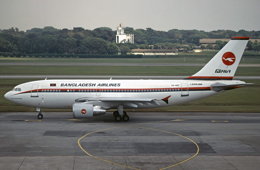 A Biman Bangladesh Airlines Airbus plane that has come to a a stop on a concrete runway. There is green grass and foliage in the background of the photo, alongside some buildings. The sky is grey and cloudy.