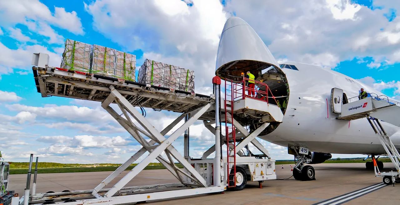 Picture of an open freight aircraft with an employee loading lots of wooden crates into its fuselage. The cargo is raised by lift equipment, keeping it aligned with the freight plane's nose. The sky is bright blue with a scattering of clouds.