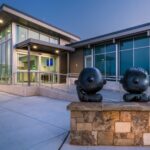 Two dark bronze statues of cartoon characters sit on a stone wall outside a modern glass-fronted building with a ramp entrance at twilight.