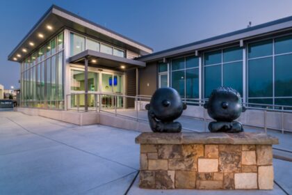 Two dark bronze statues of cartoon characters sit on a stone wall outside a modern glass-fronted building with a ramp entrance at twilight.