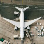 A birds eye view of a white aircraft parked on the tarmac at an airport gate surrounded by cars.