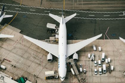 A birds eye view of a white aircraft parked on the tarmac at an airport gate surrounded by cars.
