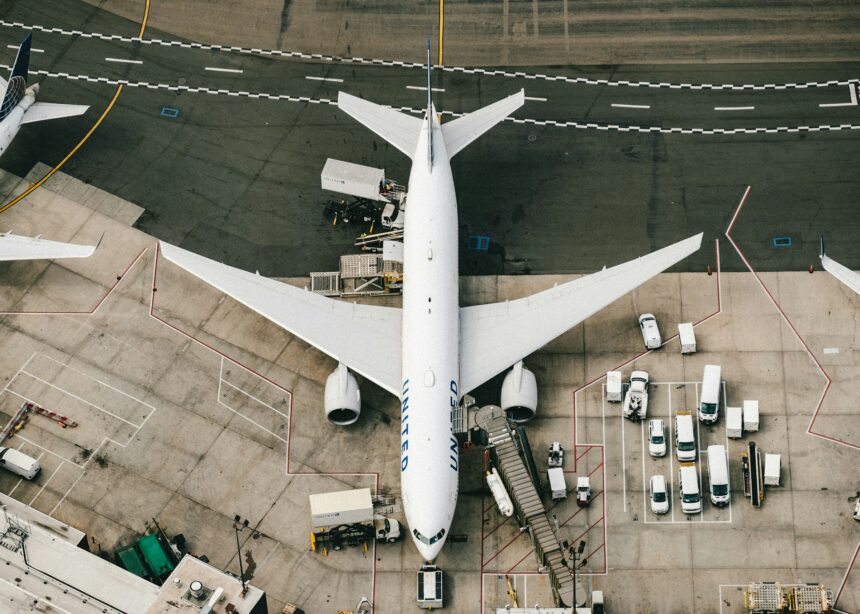 A birds eye view of a white aircraft parked on the tarmac at an airport gate surrounded by cars.