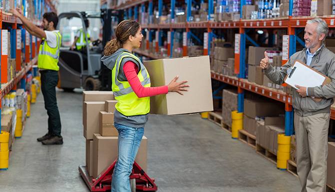 Woman in a hi-vis vest holds a large cardboard box beside a loaded pallet while a man with a clipboard gives a thumbs-up in a warehouse aisle.