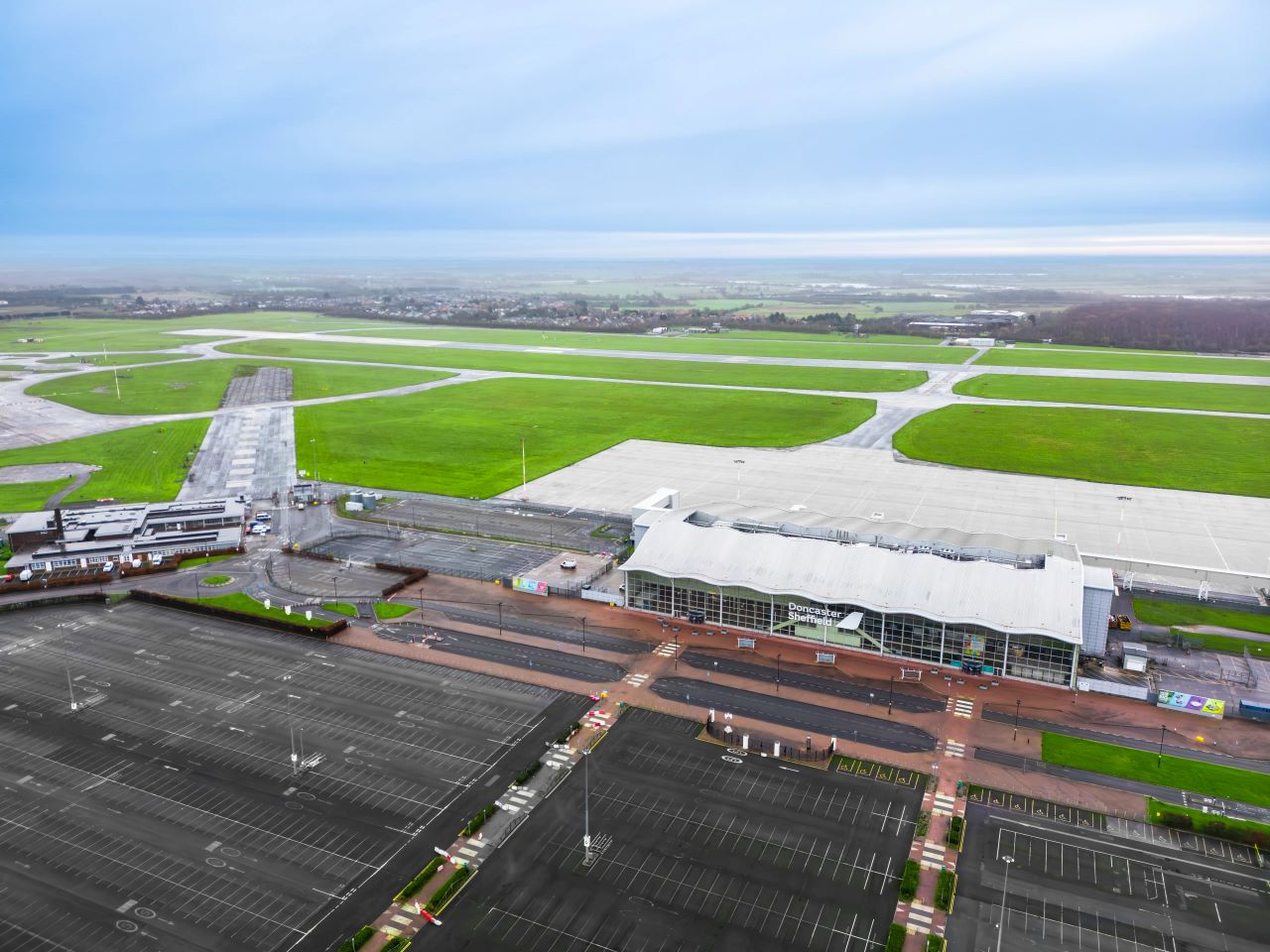 An aerial image of Doncaster Sheffield Airport depicting the empty runway and car park. Despite its emptiness, the airport looks well-maintained, with lush green grass and fresh-looking tarmac. The airport's main building is white with glass panelling and has its name written out.