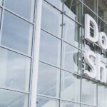 An up-close image of the outside of the entrance of Doncaster Sheffield Airport. The building is made of glass panels which reflect the sky and clouds. Viewable is white lettering that says 'Doncaster Sheffield'.